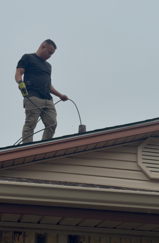 Technician on a roof peak feeding a cable into a vent pipe with a yellow drill